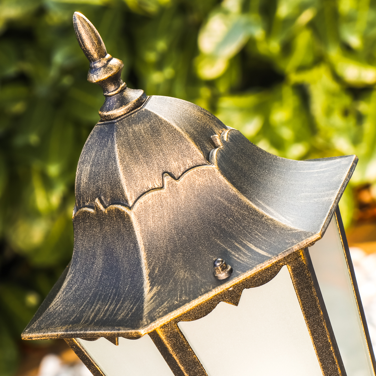 Nahaufnahme der Oberseite einer dekorativen Außenlampe. Die Lampe hat eine gold-schwarze Oberfläche und steht vor Grün.