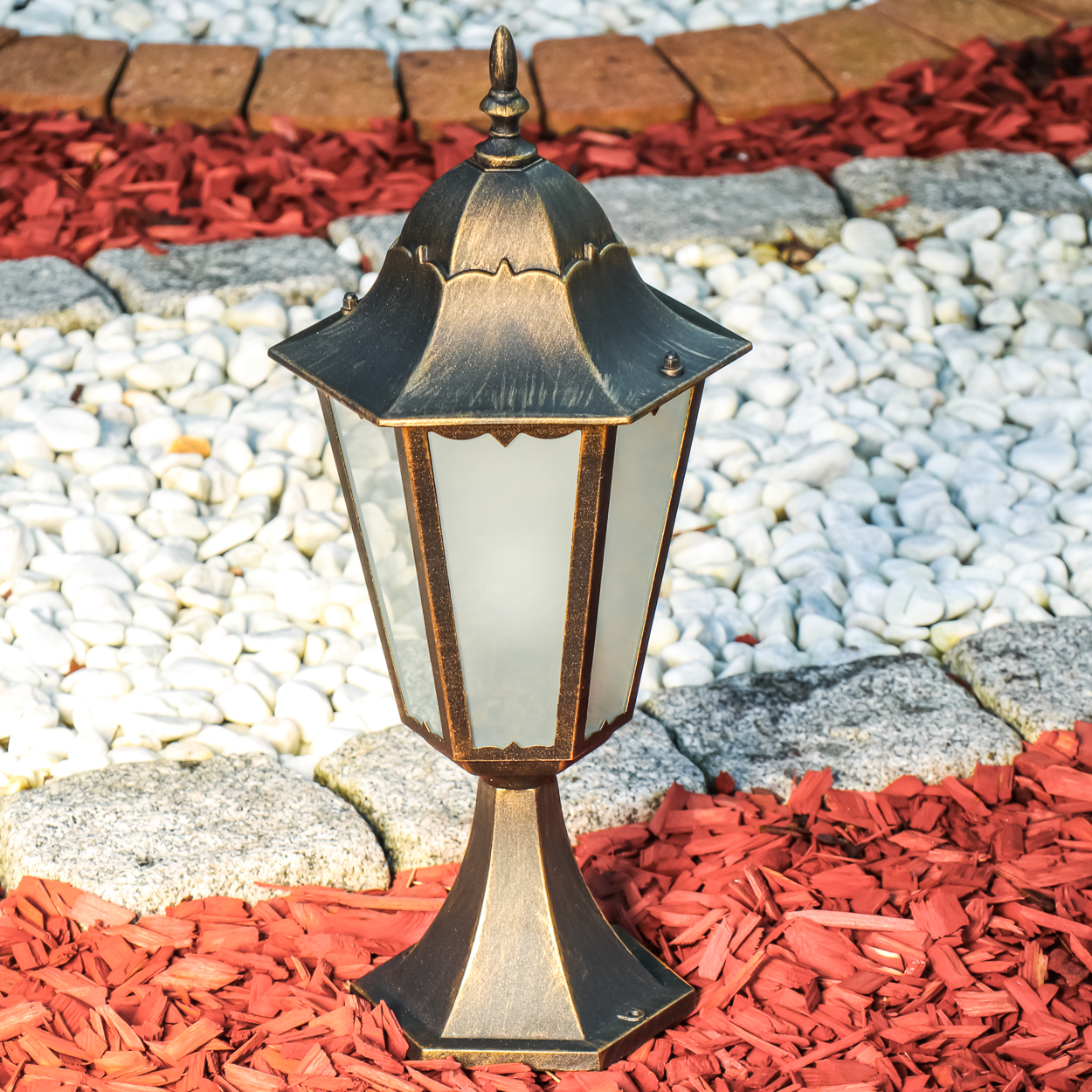 Eine dekorative Außenlampe auf einem Beet aus roter Mulch und weißen Steinen. Die Lampe hat eine gold-schwarze Oberfläche.