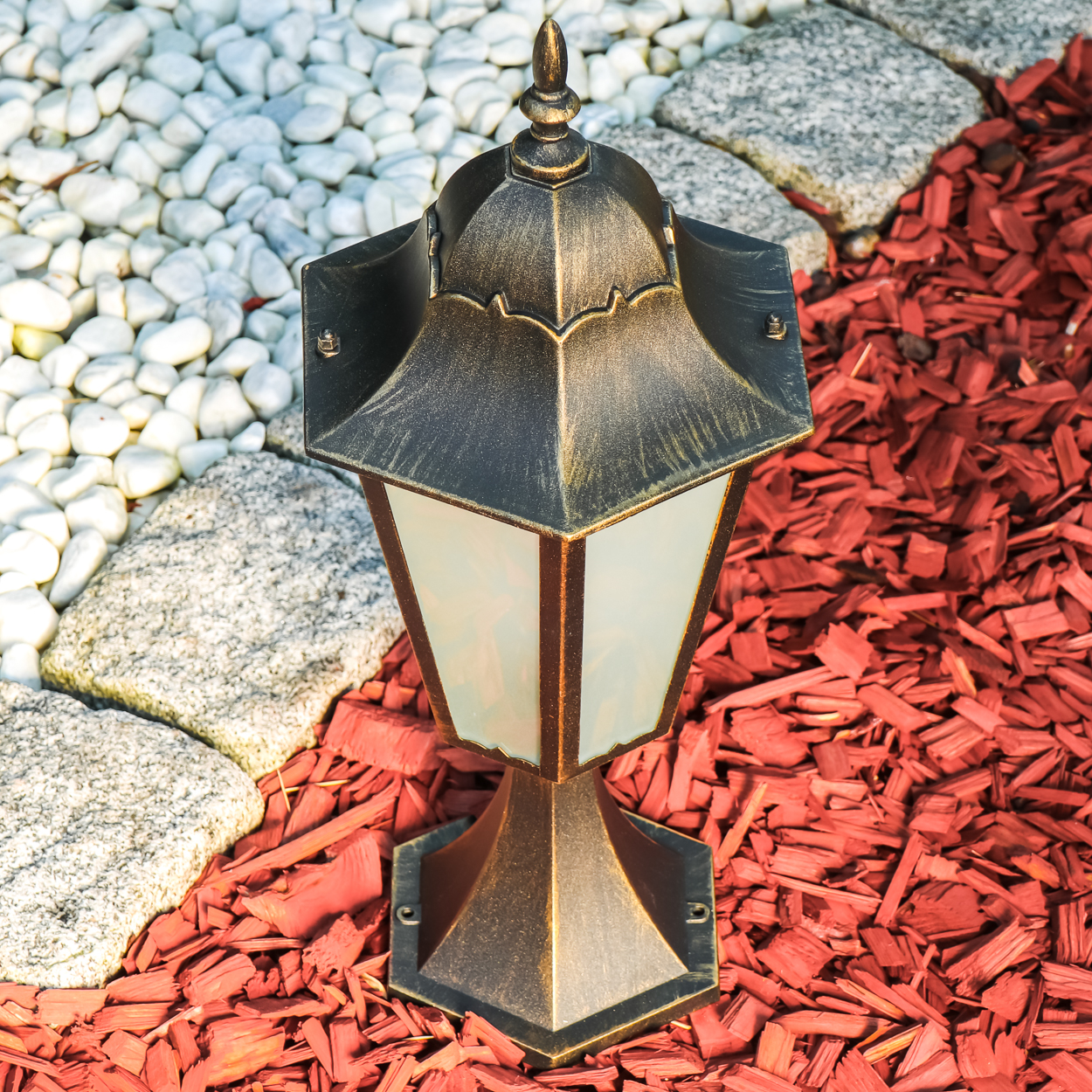 Eine dekorative Außenlampe auf einem Beet aus roter Mulch, weißen Steinen und einem Steinrand. Die Lampe ist gold-schwarz.