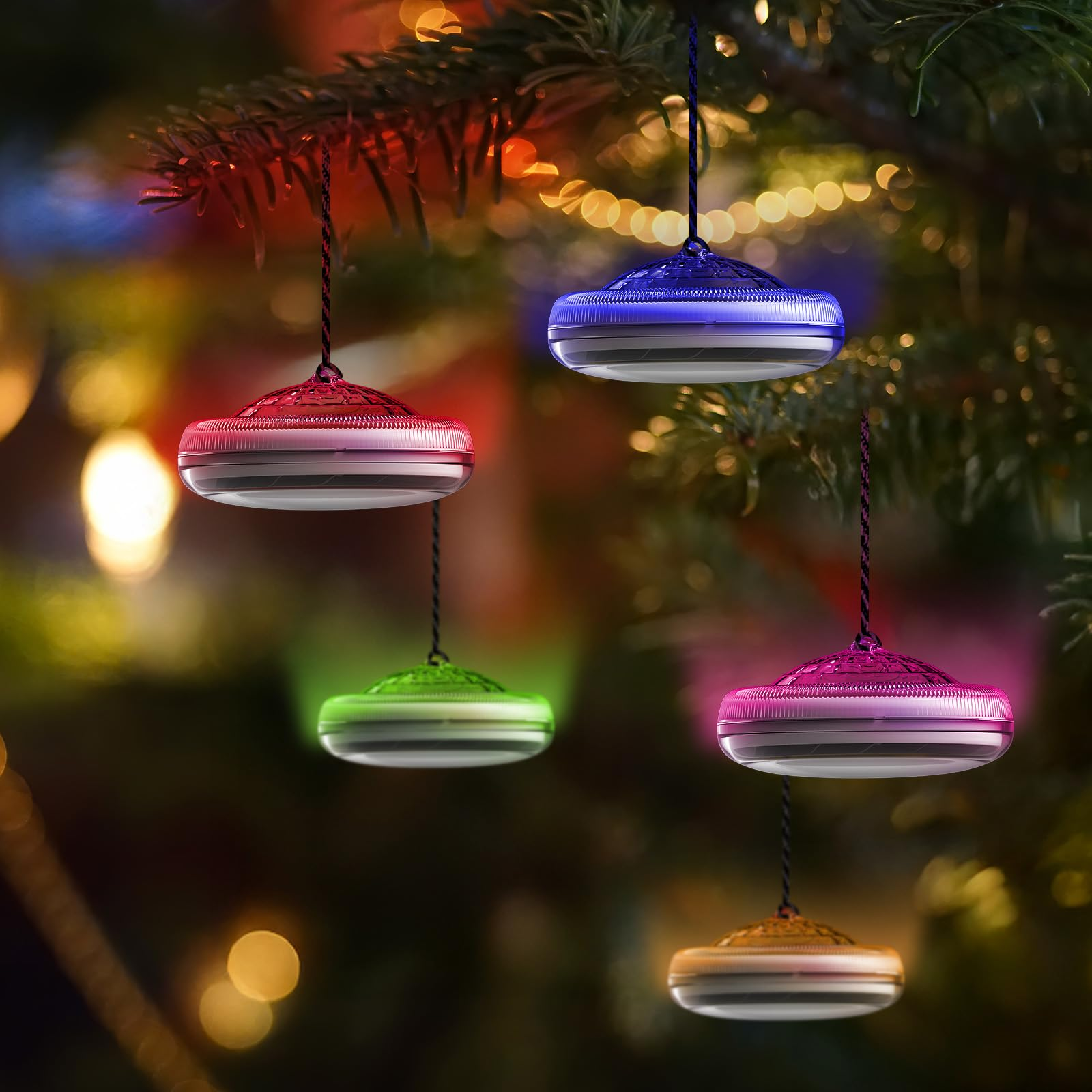 Vier kleine, bunte Hängelampen an einem Weihnachtsbaum. Jede Lampe hat eine andere Farbe: rot, grün, rosa und gold.