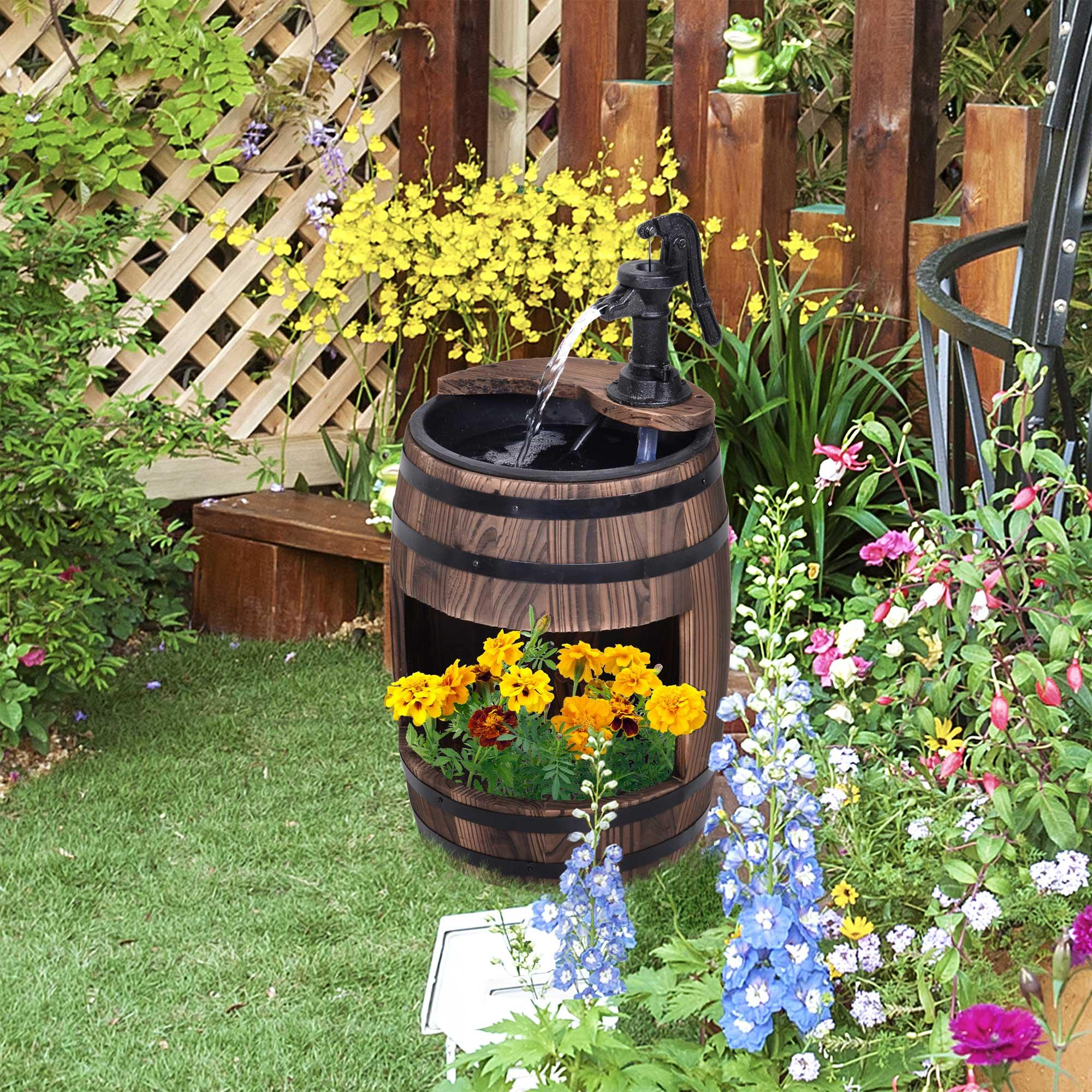 Holzfassbrunnen mit schwarzer Pumpe, Wasser fließt in das Fass, umgeben von Blumen und Grün.