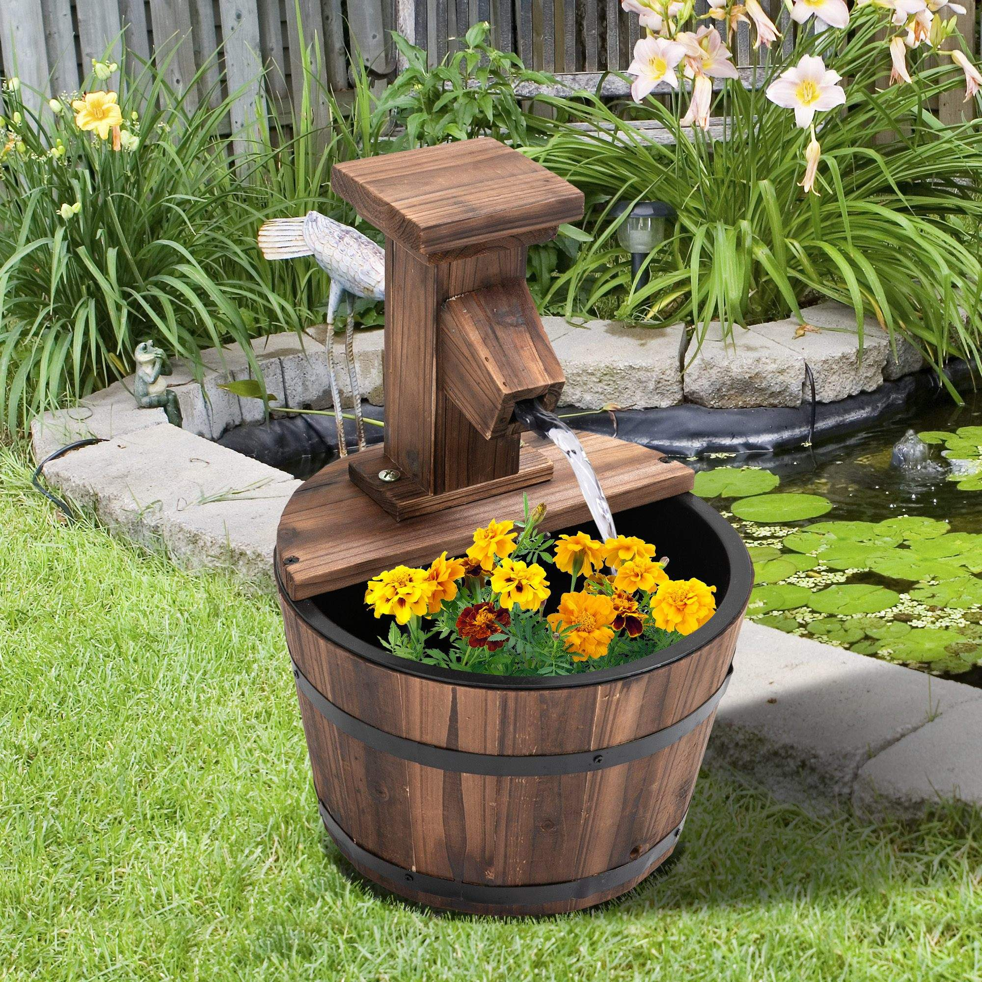 Holzbrunnen mit Wasser, das in ein Fass fließt, Blumen im Vordergrund, Grün und ein Teich im Hintergrund.