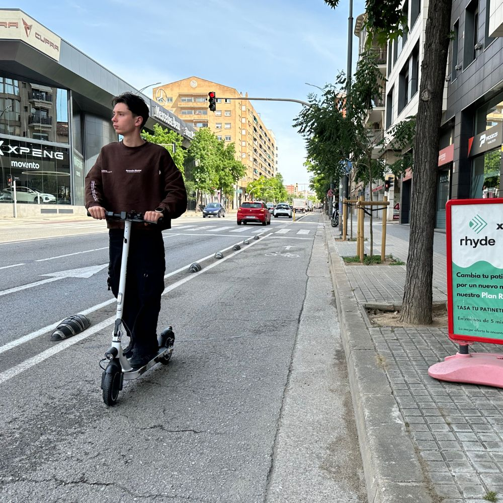 Un hombre montando un patinete blanco por una calle. Edificios y árboles a ambos lados.