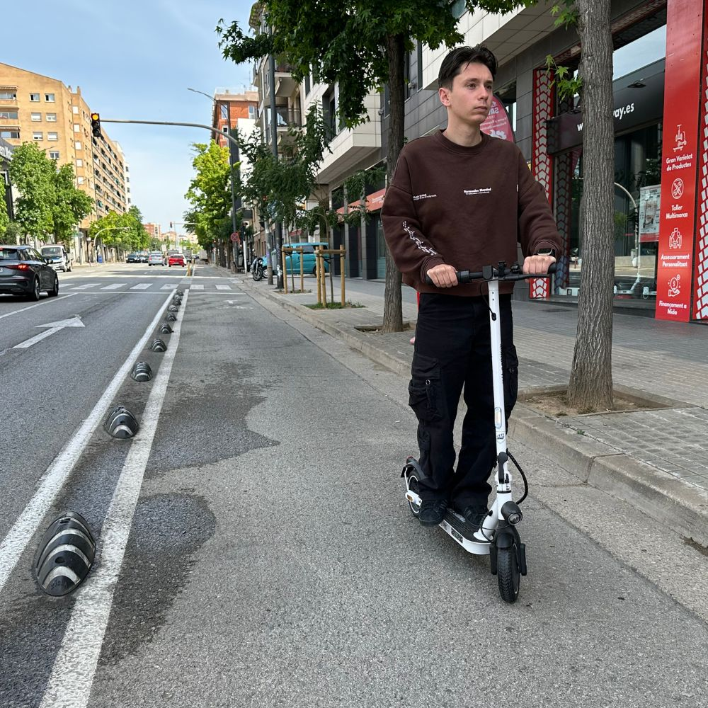 Un hombre monta un patinete blanco en una calle. Edificios y árboles a ambos lados. Cielo azul al fondo.