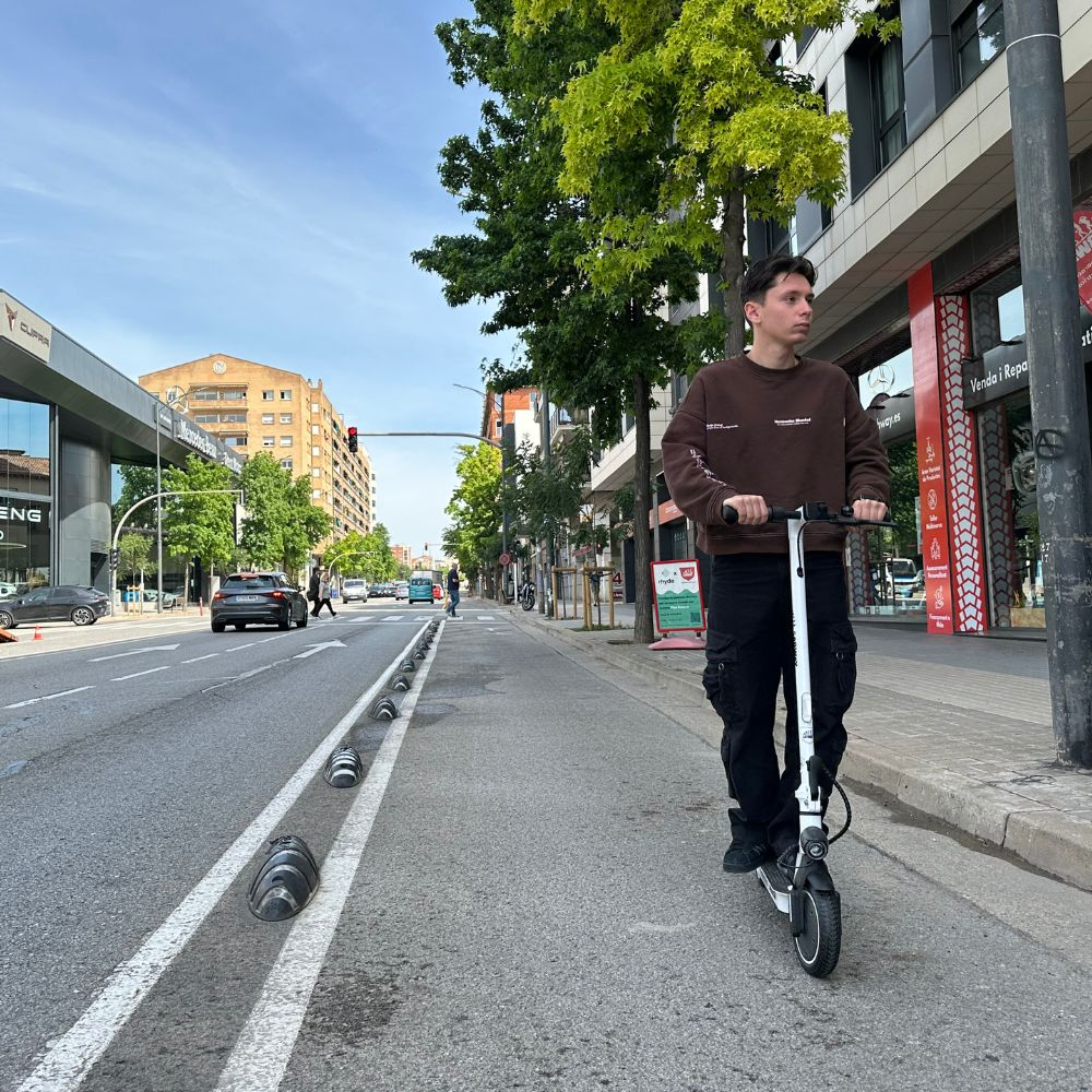 Un hombre monta un patinete blanco en una calle. Edificios y árboles al fondo.