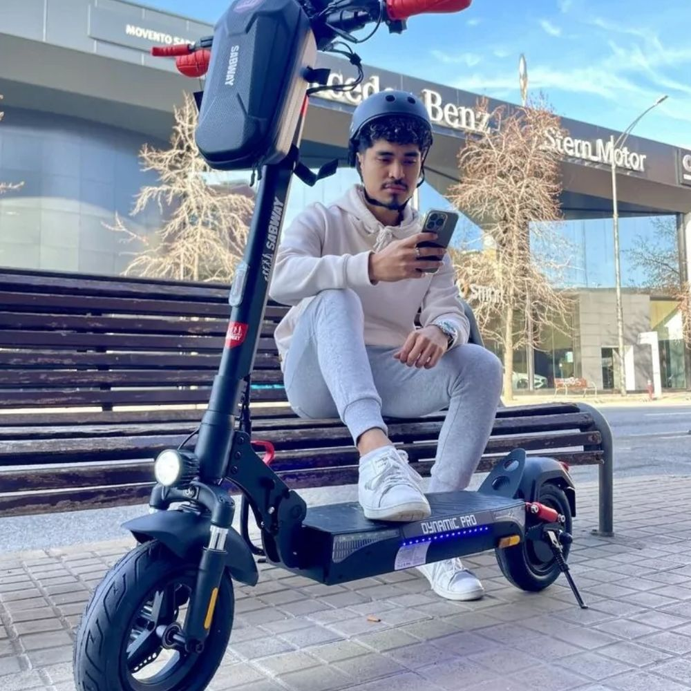 Un hombre con sudadera blanca y pantalones grises sentado en un patinete eléctrico negro, sosteniendo un teléfono.