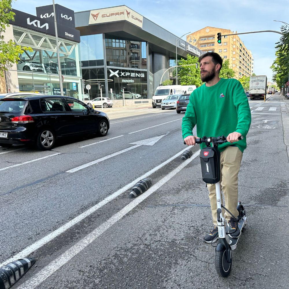 Un hombre con un suéter verde montando un patinete eléctrico blanco y negro frente a un edificio.