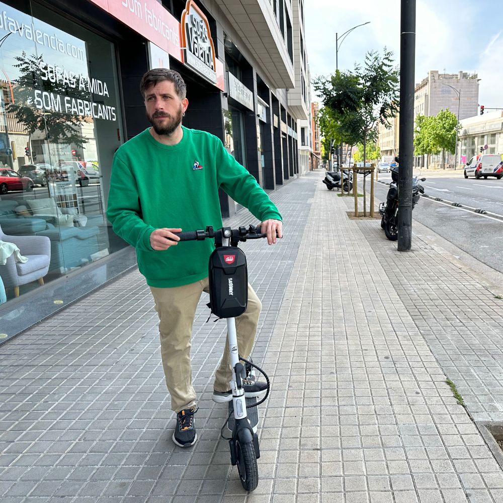 Un hombre con un suéter verde monta un patinete eléctrico blanco y negro en una acera de la ciudad.