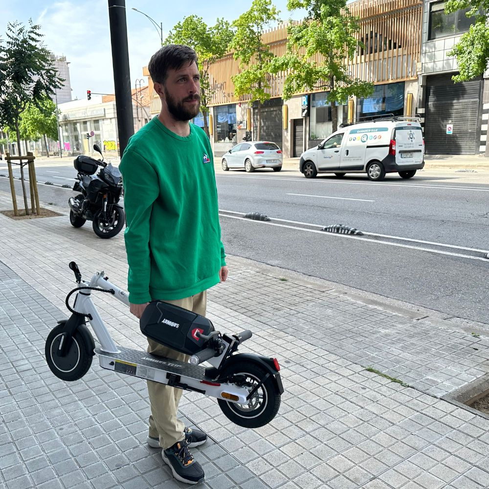 Un hombre de verde sostiene un patinete blanco en la acera. Una moto y vehículos están en el fondo.