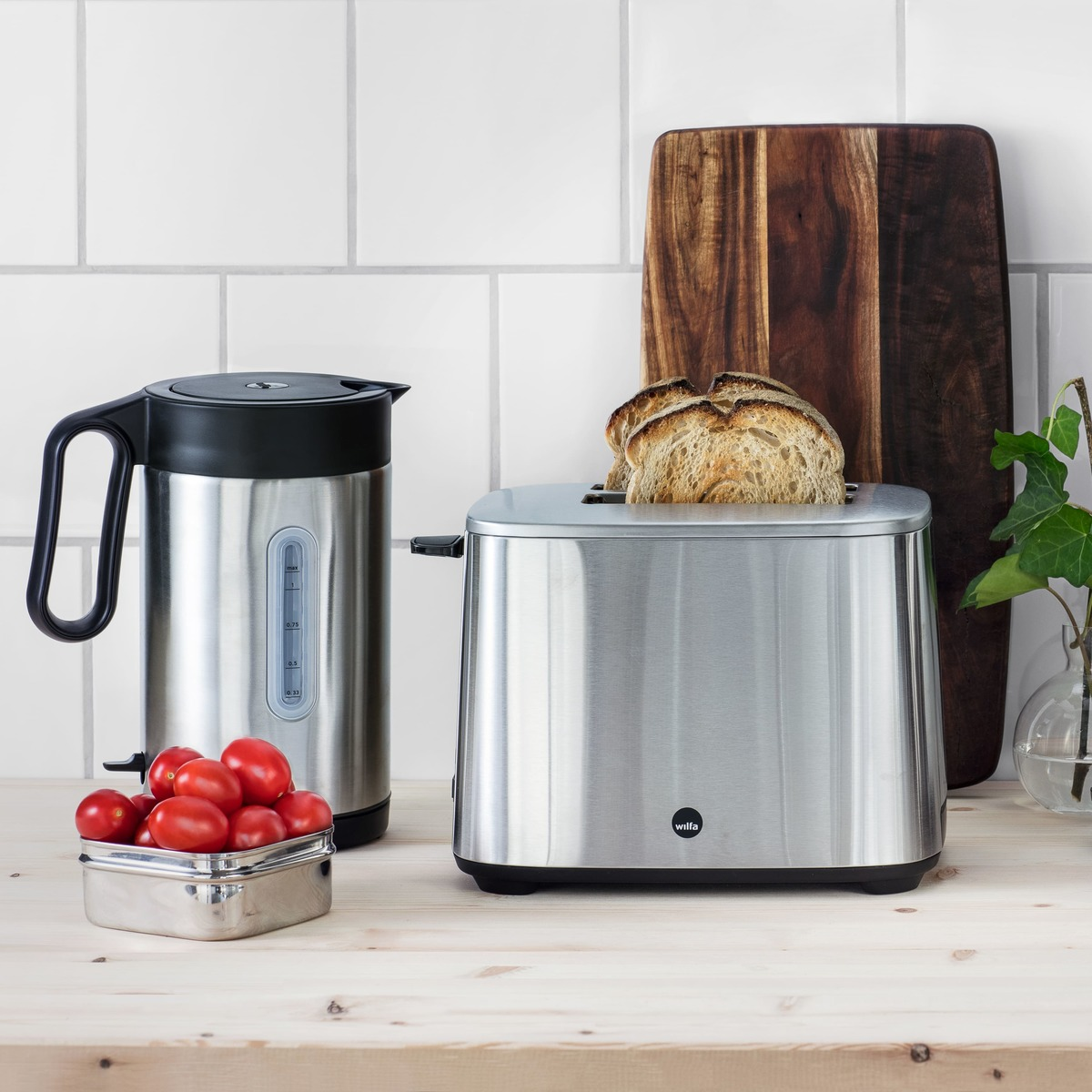 Toaster, Wasserkocher, Tomaten und Brot auf einer Holztheke. Grüne Pflanze sichtbar.