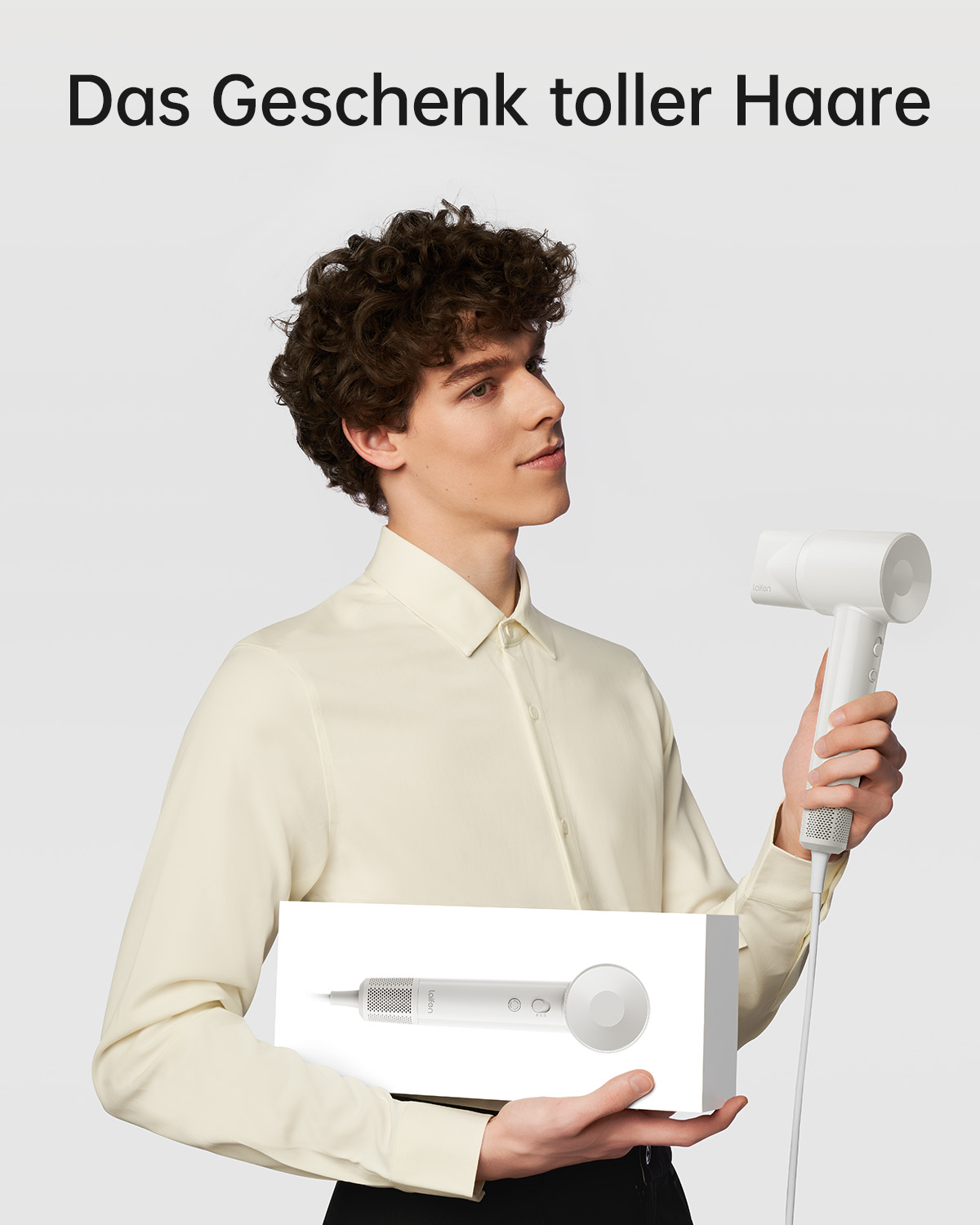 Ein Mann in einem hellen Hemd hält einen weißen Haartrockner und eine weiße Schachtel mit einer Haartrocknerabbildung.