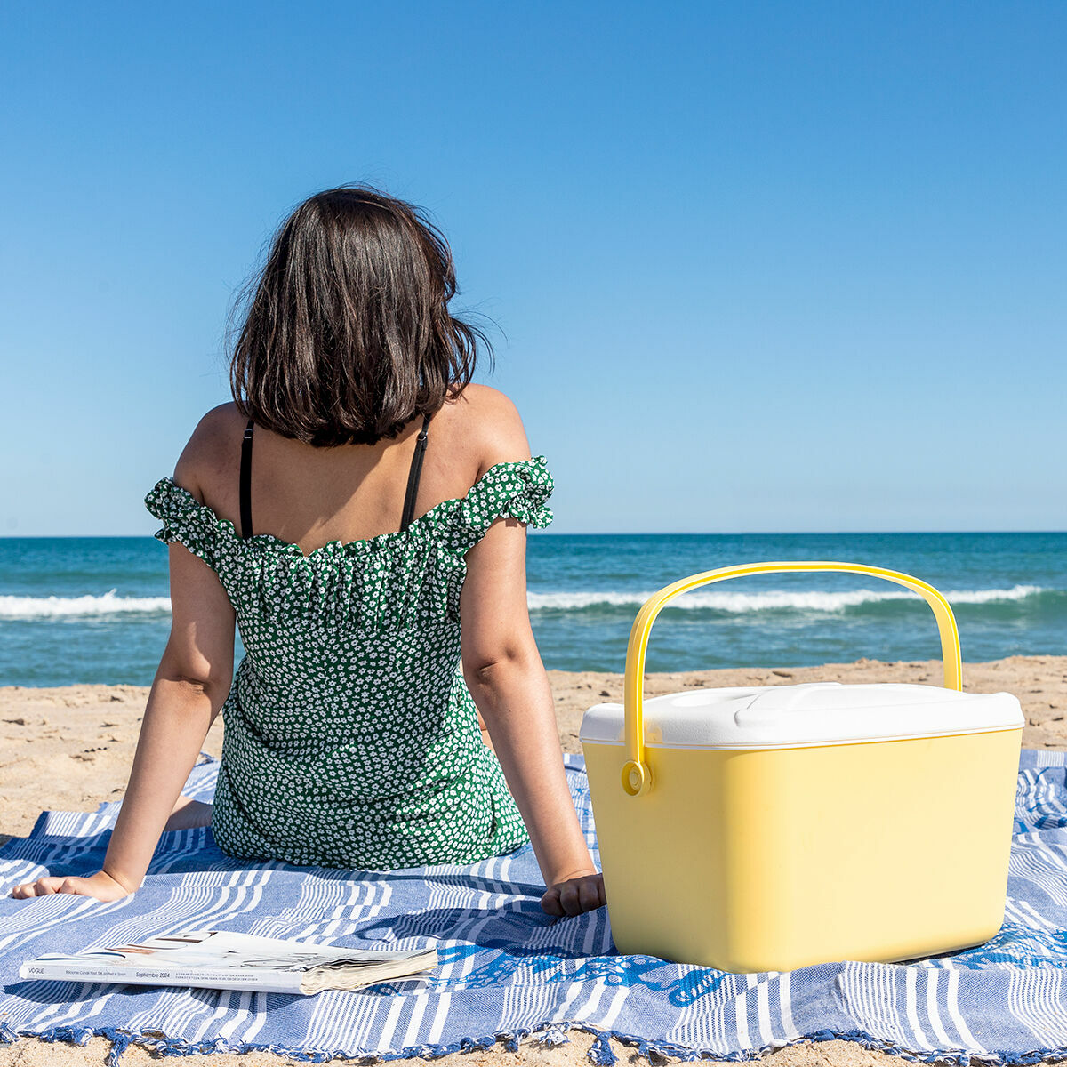 Una mujer sentada en la playa, mirando al mar, junto a una nevera amarilla.