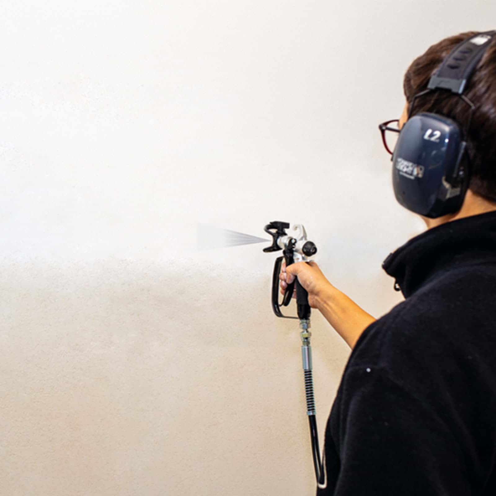 Eine Person mit Kopfhörern und Brille benutzt eine Spritzpistole, um eine weiße Wand in einem gut beleuchteten Raum zu streichen.