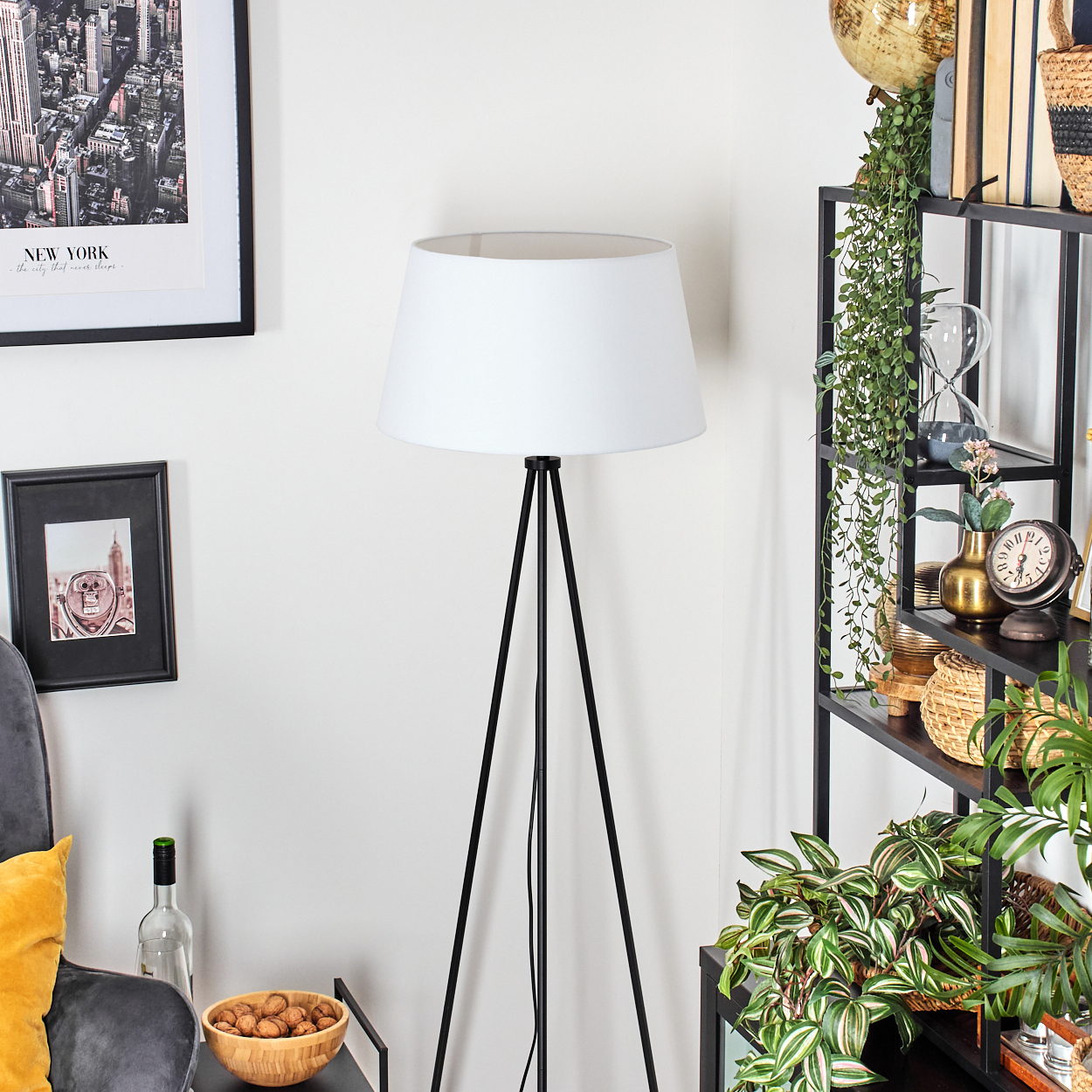 Eine Stehlampe mit weißem Lampenschirm steht in einem Raum mit Bücherregal, Globus und Pflanzen.