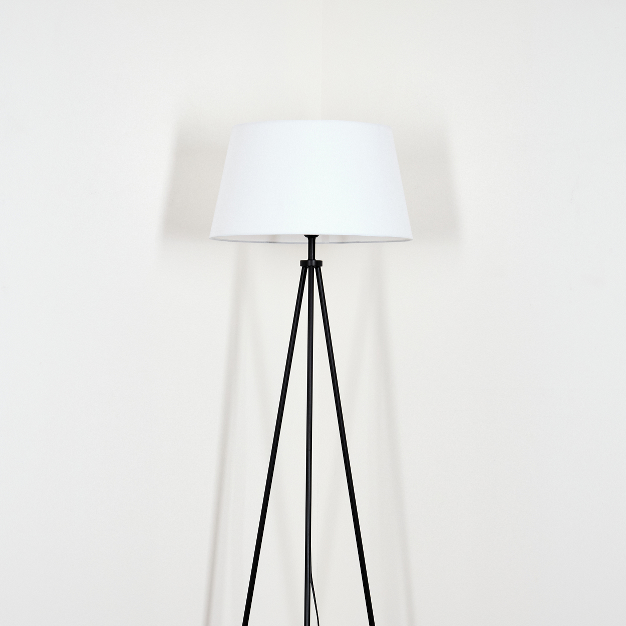 Eine Stehlampe mit weißem Lampenschirm und schwarzen Stativbeinen. Sie steht in einer Zimmerecke.