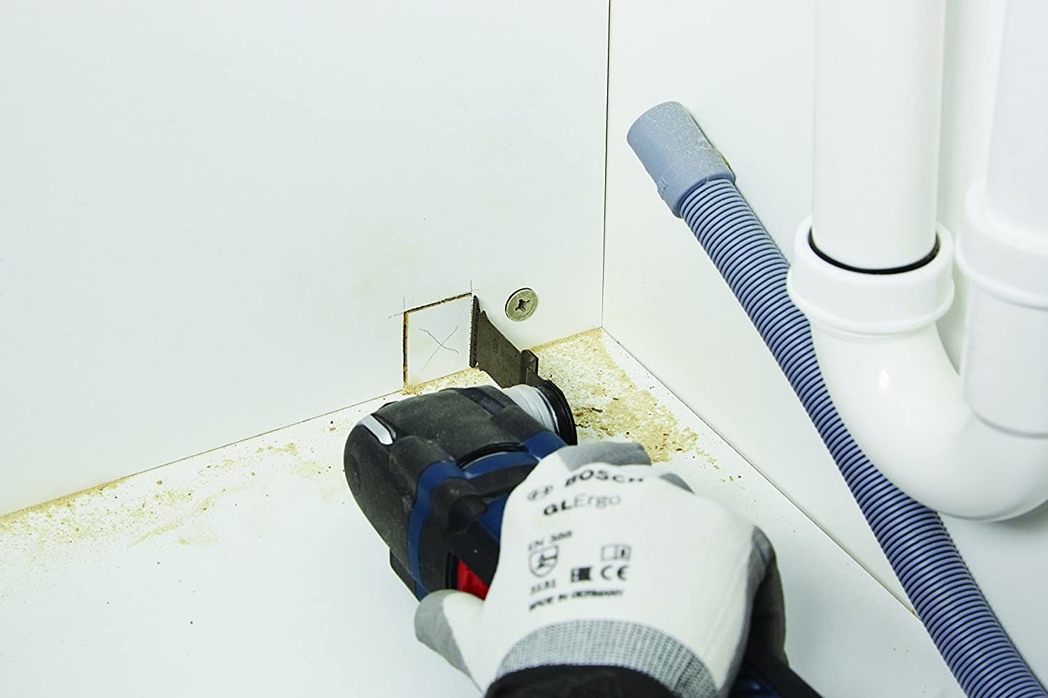 Ein Bosch Multi-Tool schneidet in eine Ecke eines weißen Schranks. Ein graues Rohr ist sichtbar.