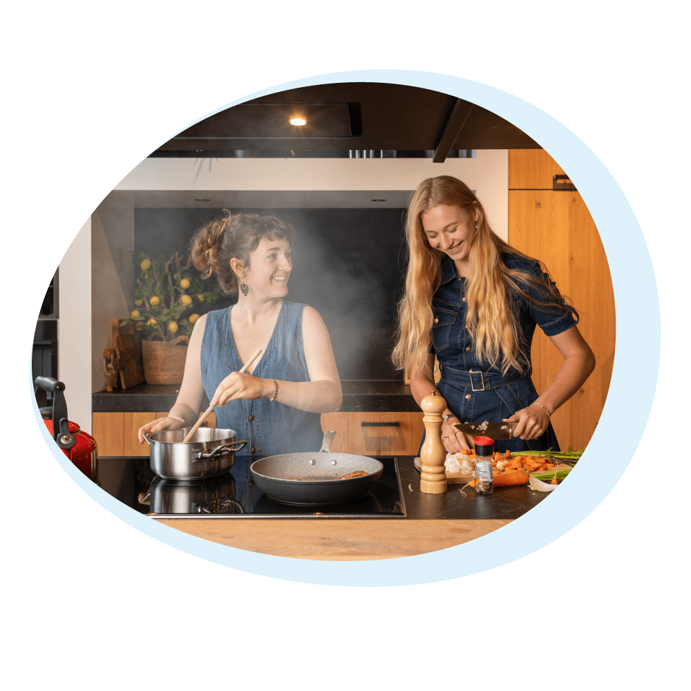 Due donne cucinano in cucina. Una mescola in una pentola, l'altra taglia le verdure. La scena è luminosa.