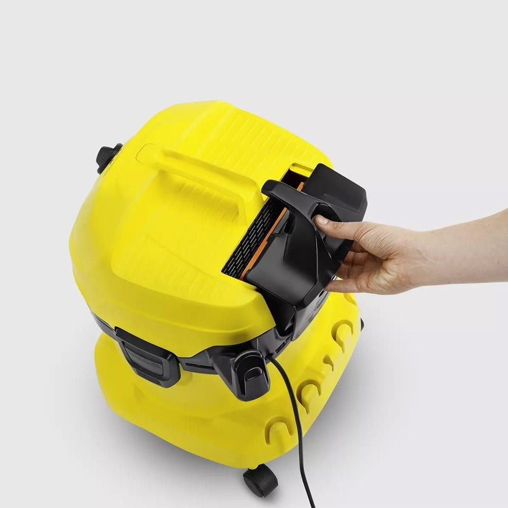 Aspirapolvere giallo con una mano che solleva un coperchio nero. Sfondo bianco. Cavo di alimentazione nero.