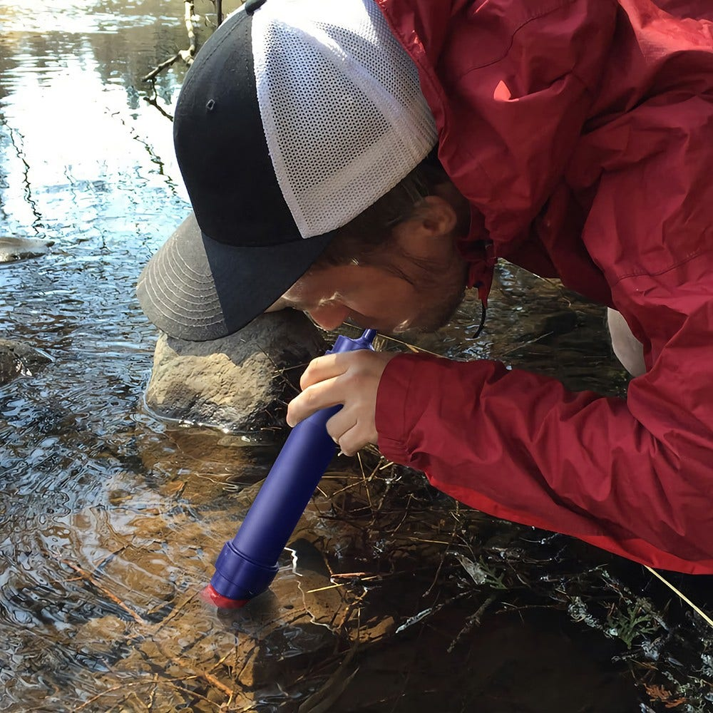 Una donna con giacca rossa e cappello filtra l'acqua da un ruscello con un filtro blu.