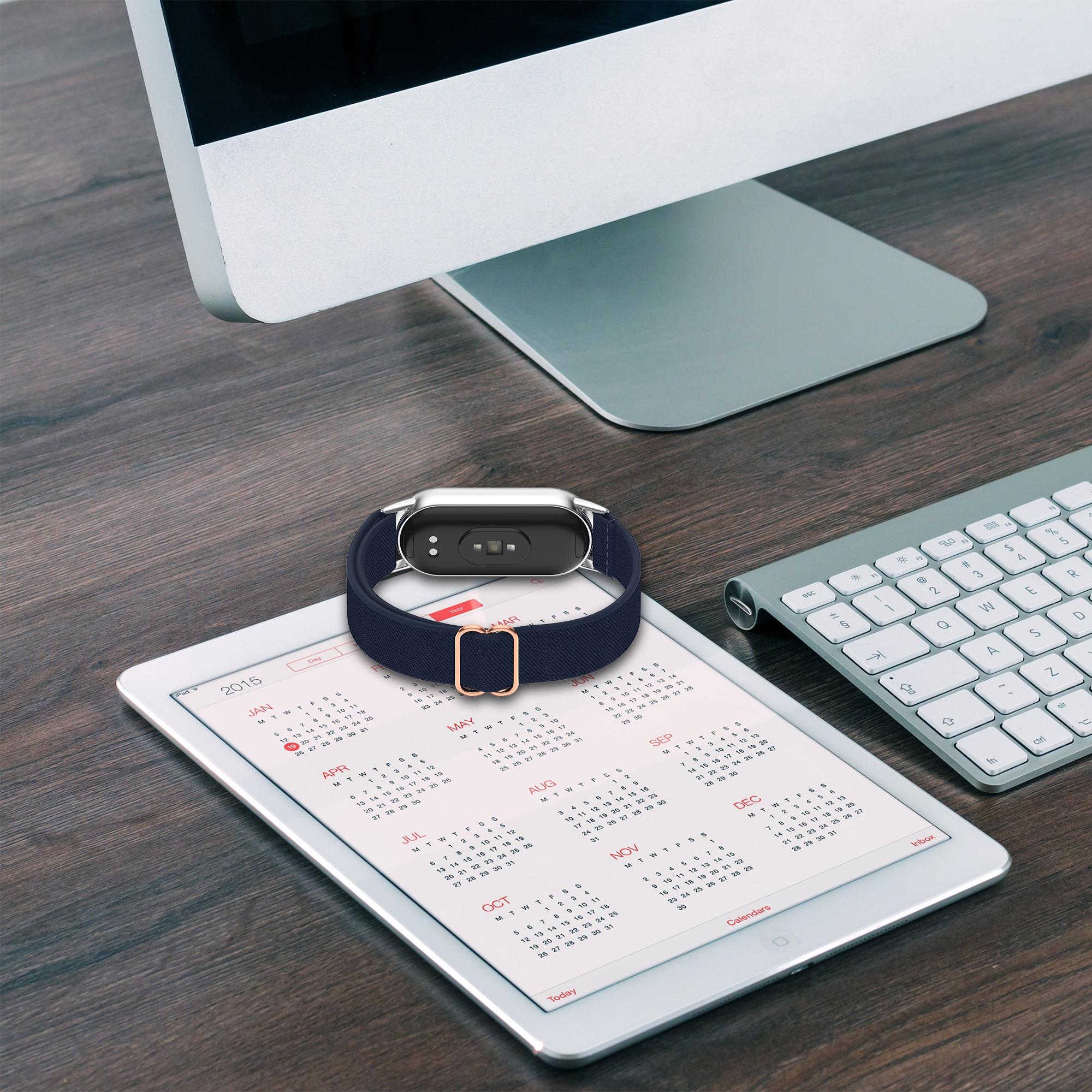 Smartwatch z ciemnoniebieskim paskiem i złotą klamrą, na otwartym kalendarzu tabletu, obok klawiatury.