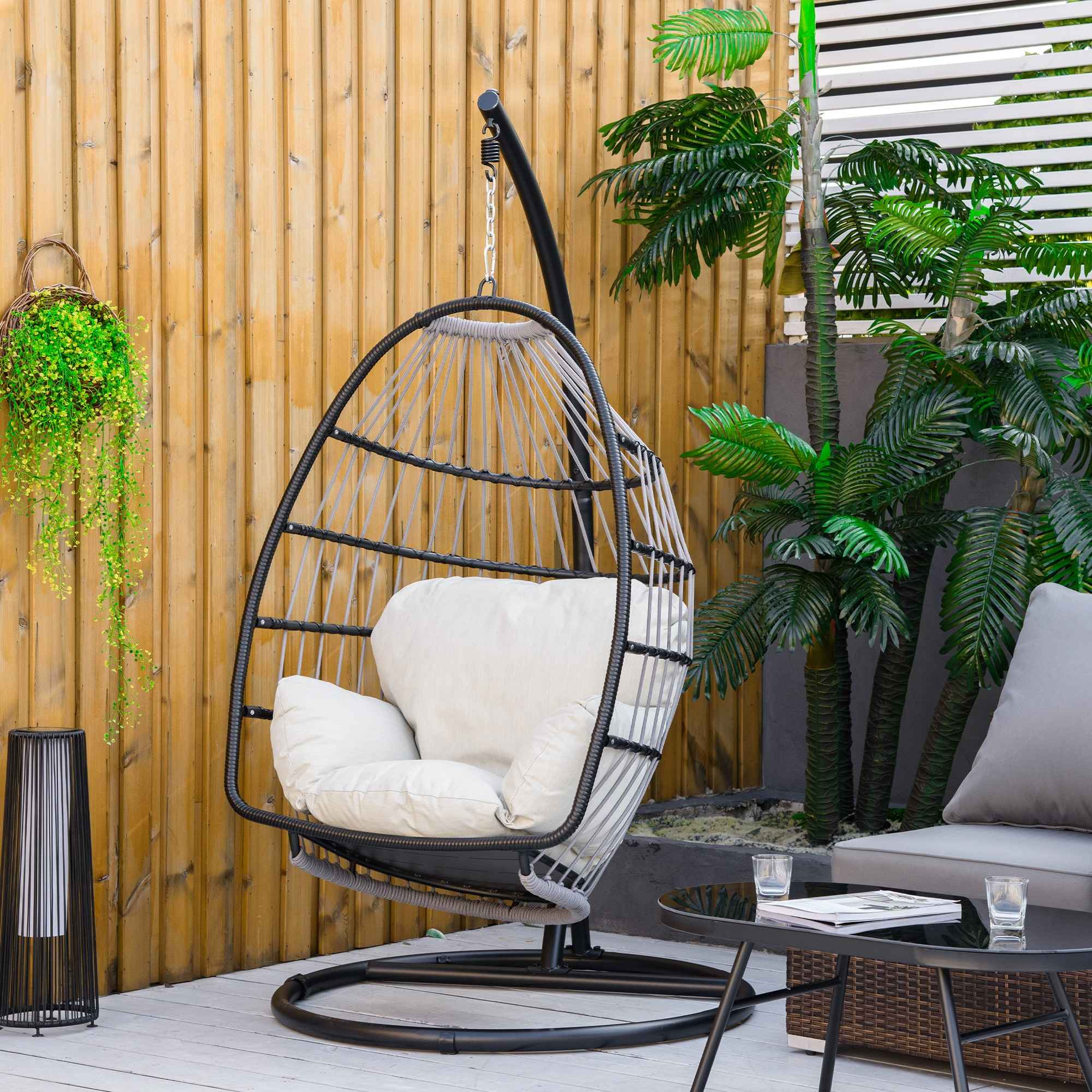 Ein hängender Eiersessel in einem Garten, mit Palmen, einem Sofa und einer schwarzen Lampe und Tisch.