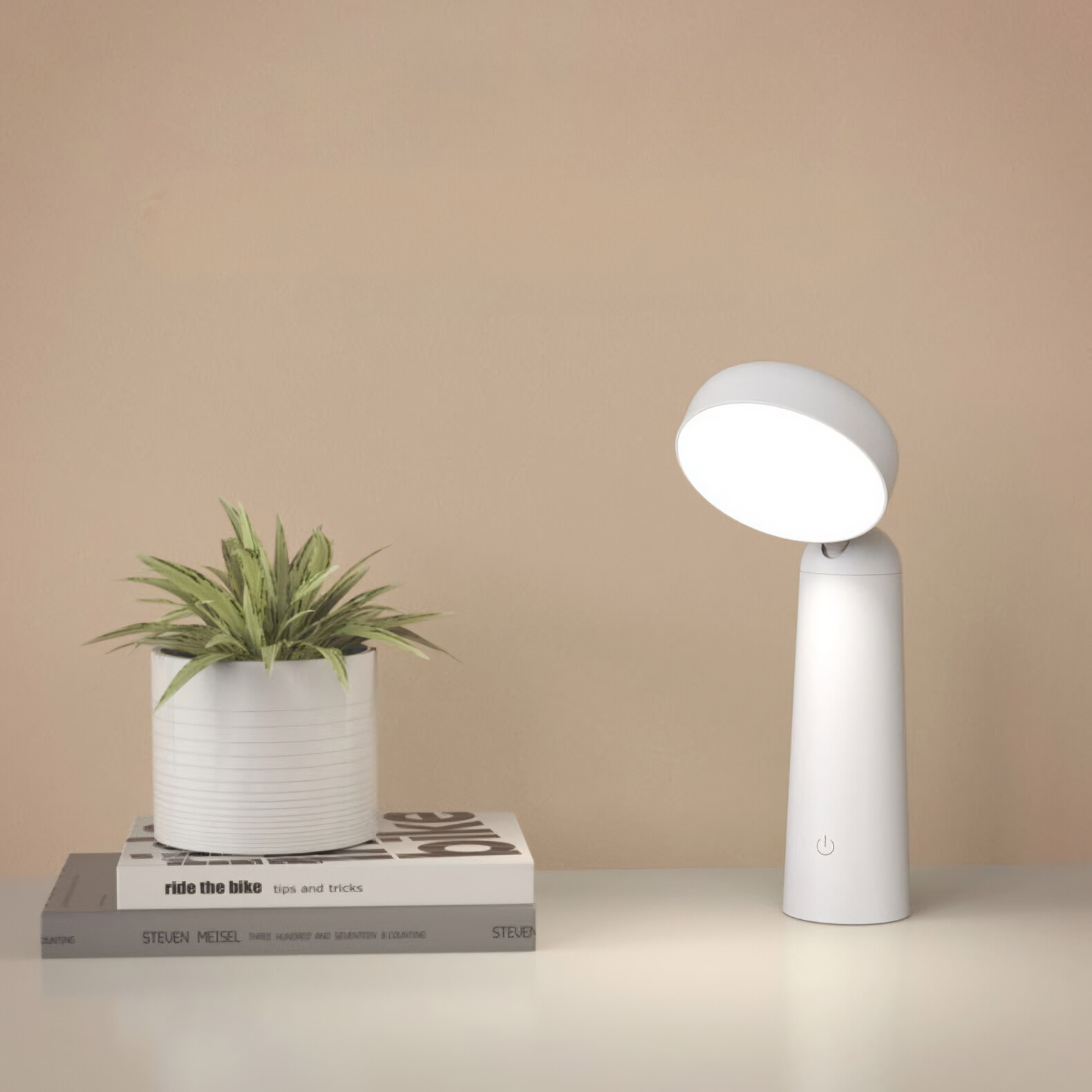 Weiße Schreibtischlampe mit rundem Licht und Pflanze im Hintergrund. Bücher auf dem Schreibtisch sichtbar.