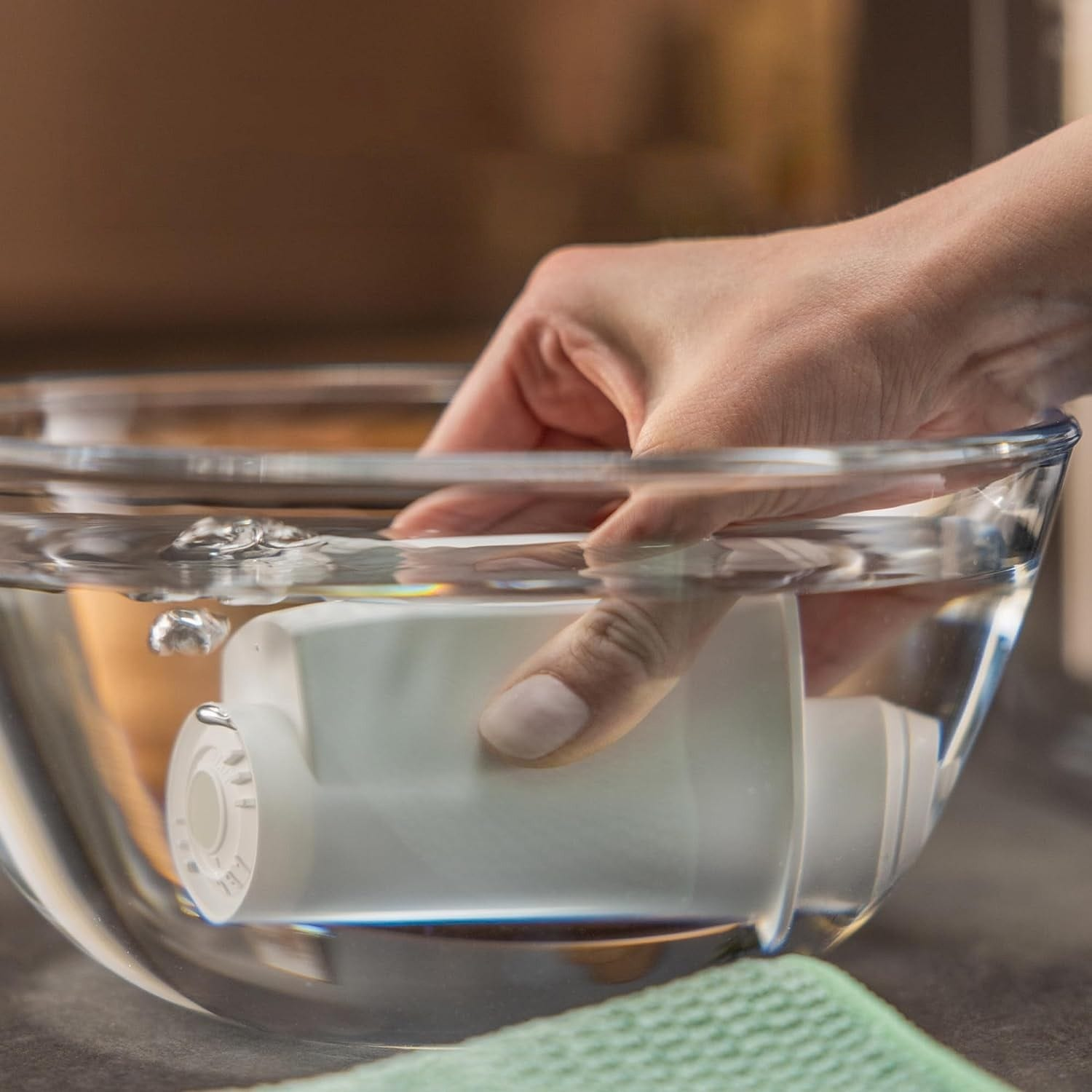 Eine Hand taucht einen weißen Wasserfilter in eine Glasschüssel mit Wasser ein.