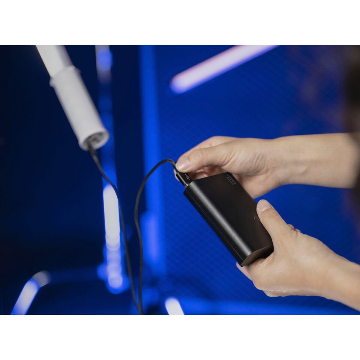 Hände verbinden ein schwarzes Kabel mit einer schwarzen Powerbank. Der Hintergrund ist blaues Neonlicht.
