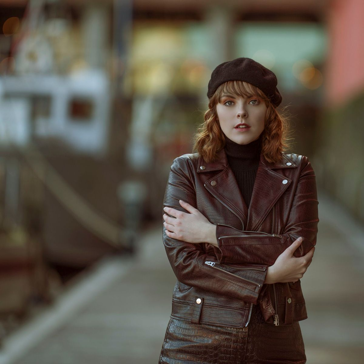 Kobieta w skórzanej kurtce i berecie stoi ze skrzyżowanymi ramionami. Rozmyte tło budynku i ścieżki.