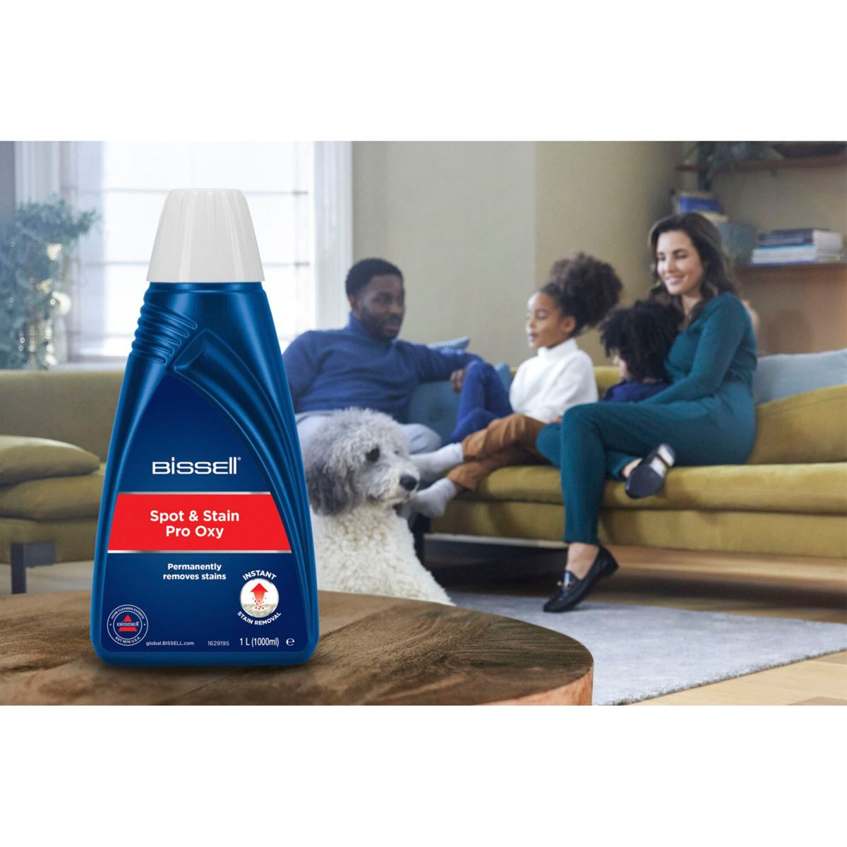 Een gezin zit op een bank en een fles vlekverwijderaar staat op de voorgrond. Er is ook een hond aanwezig.