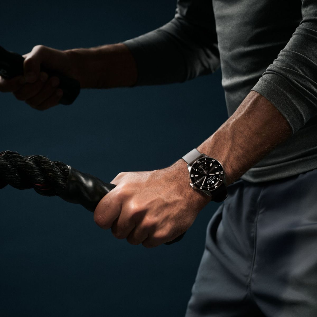 Mężczyzna trzymający czarną linę obiema rękami, noszący zegarek. Ciemne tło i szare ubrania.