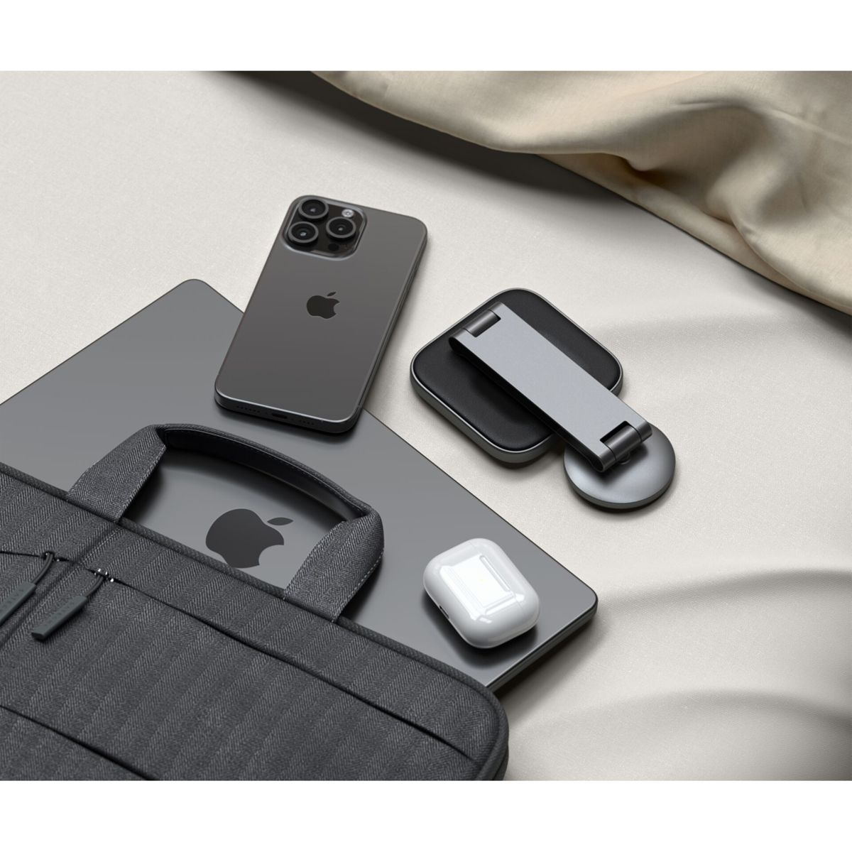 Ein Laptop, Telefon, AirPods und ein Ständer liegen auf einer Oberfläche, in der Ecke eine graue Tasche.
