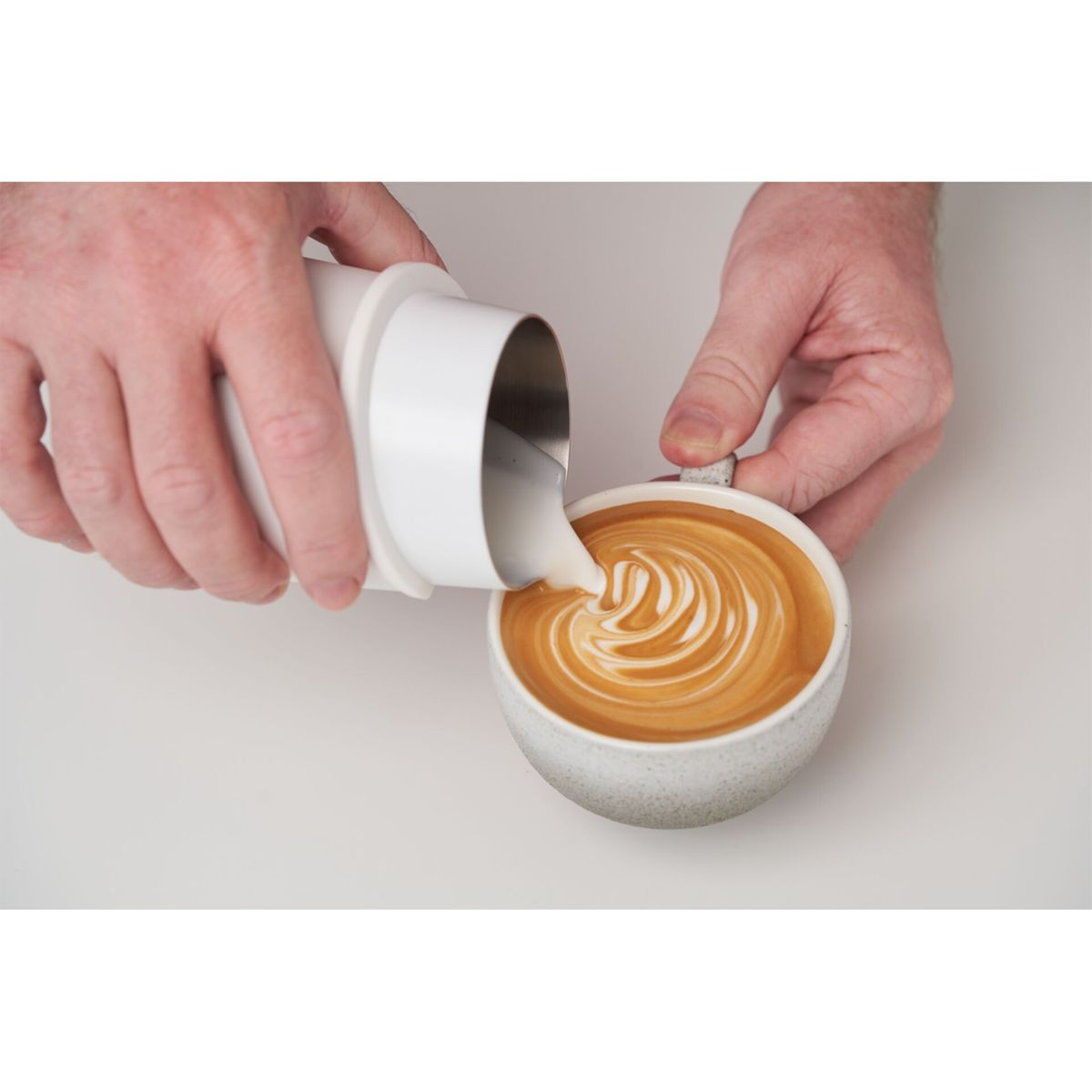 Hände gießen weiße Flüssigkeit in eine Tasse Kaffee mit weißem Design oben drauf.