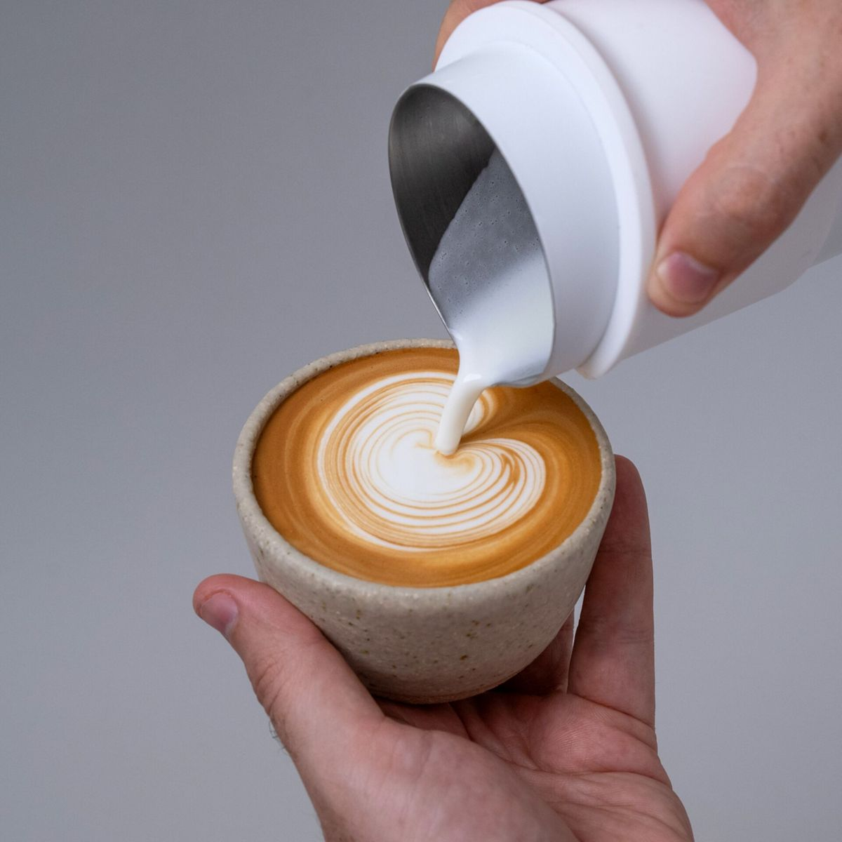 Hände gießen weiße Flüssigkeit in eine Kaffeetasse, wodurch ein herzförmiges Design entsteht.