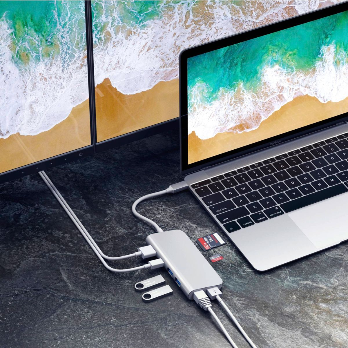 Silberner Hub an Laptop und zwei Monitoren, mit USB-Laufwerken und SD-Karte auf dunkler Oberfläche.