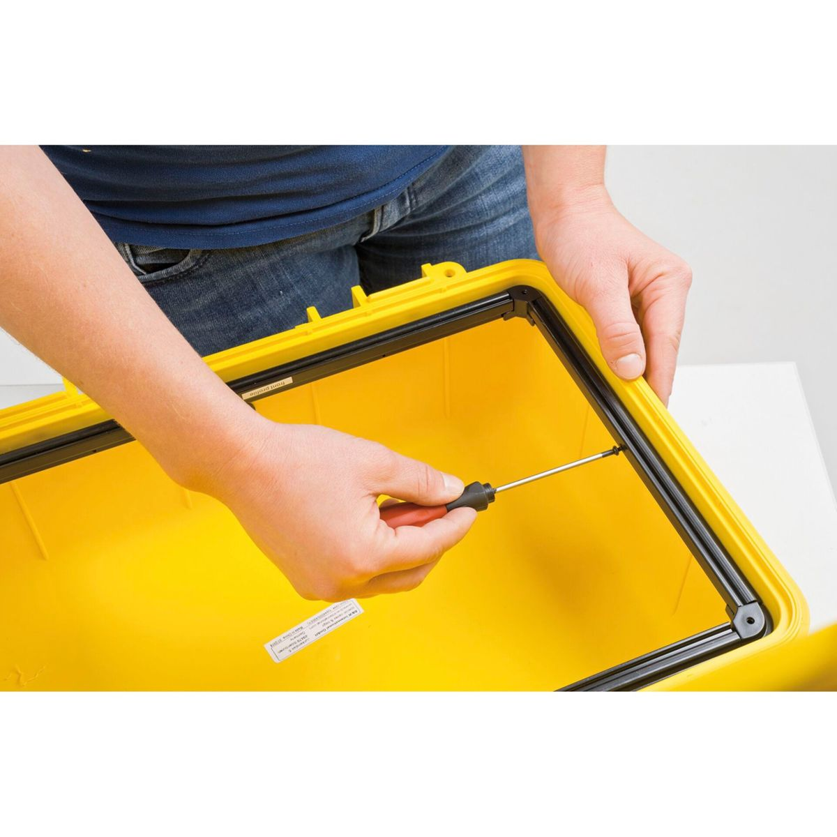 Eine Person schraubt mit einem Schraubendreher an einer gelben Box. Die Box ist offen, der Hintergrund ist weiß.