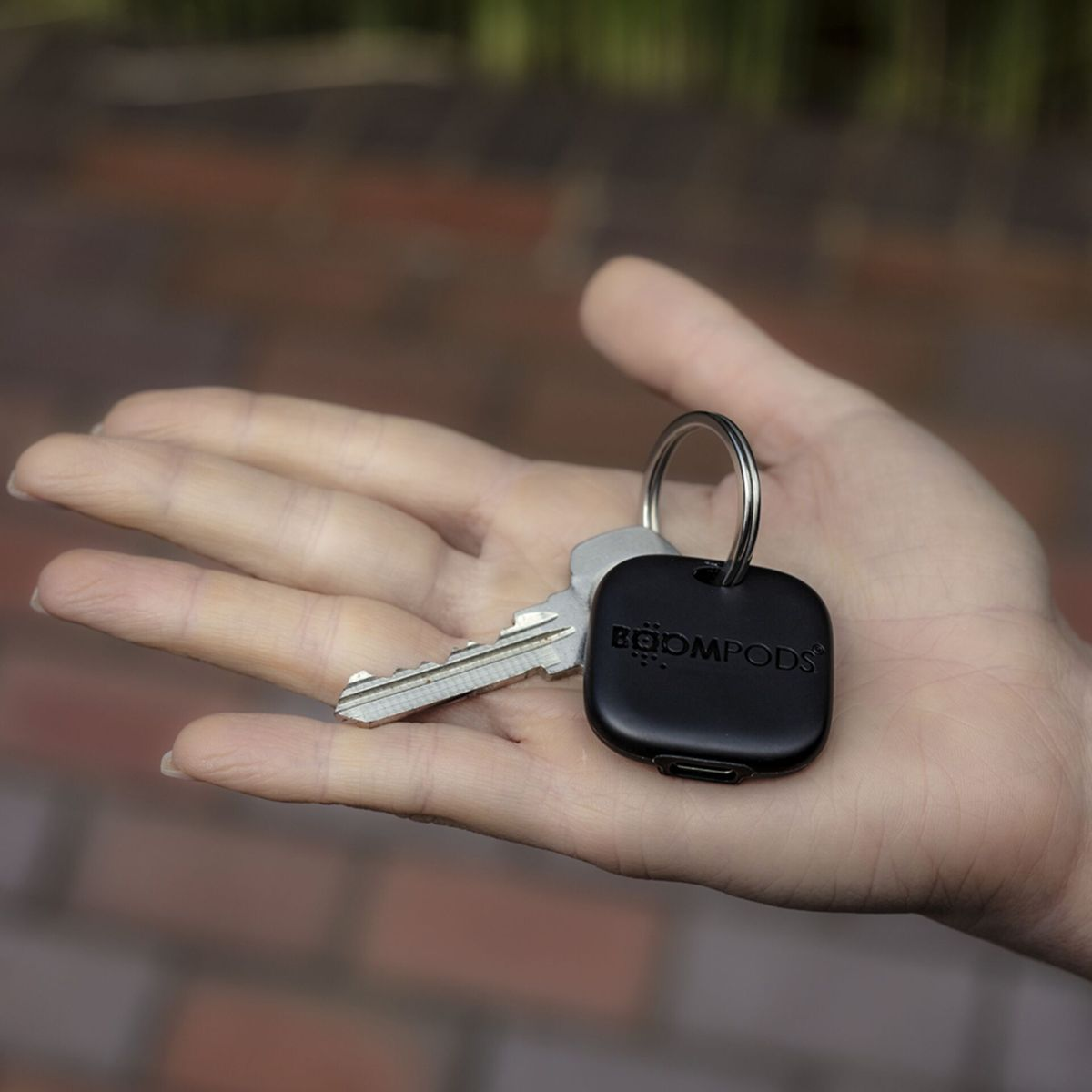 Dłoń trzyma klucze i czarny tracker Boompods. Tło to rozmyta ceglana ściana.