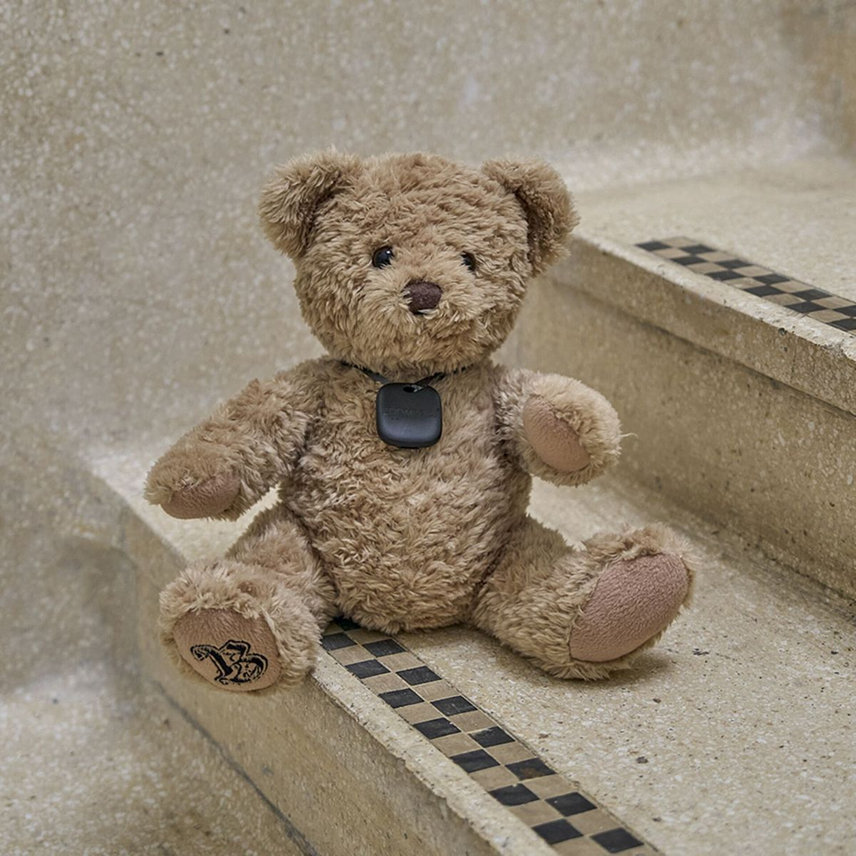 Brązowy miś siedzący na schodach z czarną zawieszką na szyi. Schody mają wzór w szachownicę.