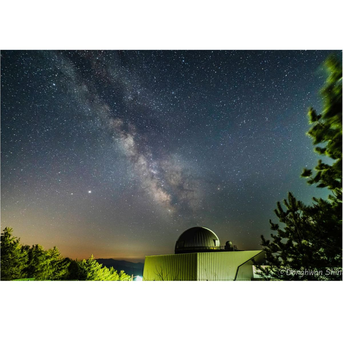 Nocne niebo z Drogą Mleczną i obserwatorium. Drzewa kadrują scenę. Niebo jest ciemne i pełne gwiazd.