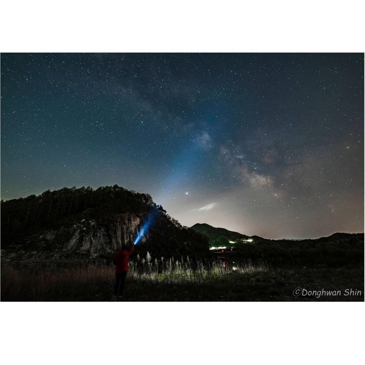 Osoba trzymająca latarkę, kierująca wiązkę światła w stronę gwiaździstego nieba nad ciemnym krajobrazem.