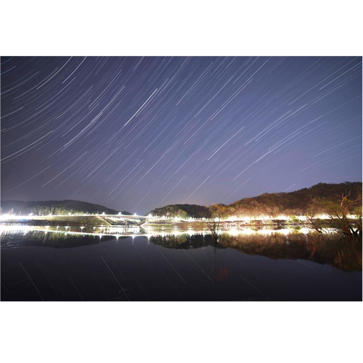 Scena nocna z jeziorem odbijającym światła mostu i gwiaździste niebo, zdjęcie z długą ekspozycją.