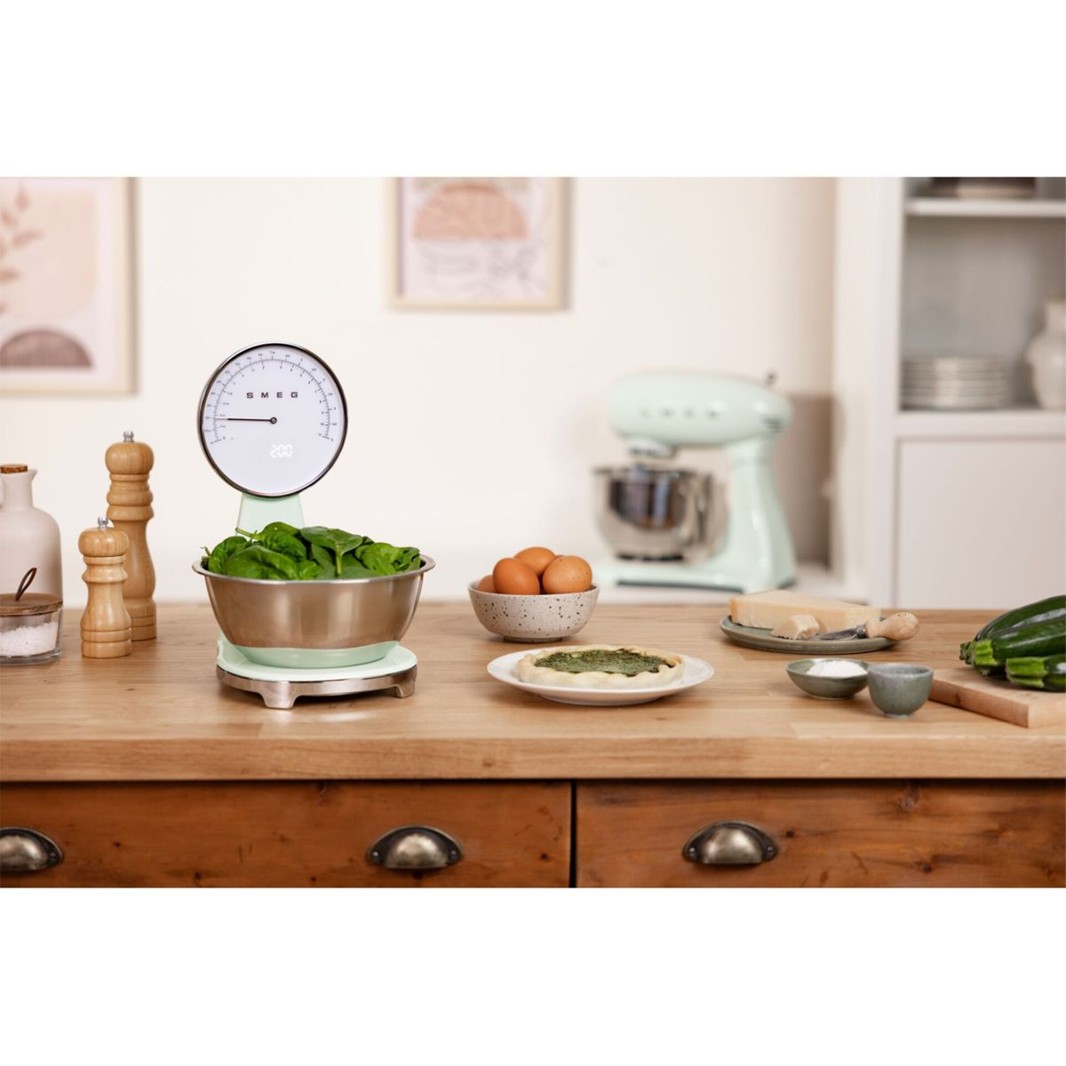 Scena kuchenna z wagą SMEG z miską ze szpinakiem. Widoczne są jajka i inne produkty spożywcze.