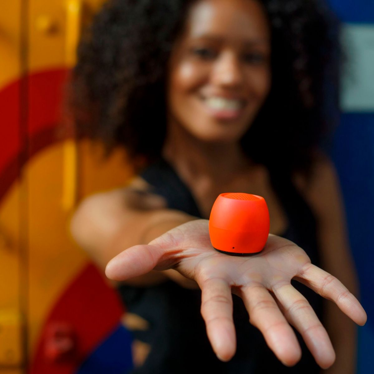 Kobieta trzymająca pomarańczowy głośnik w dłoni na tle kolorowego tła.