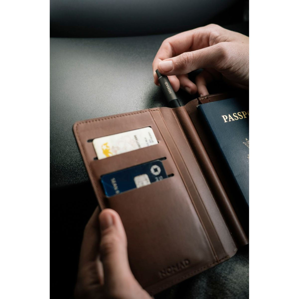 Hände halten eine braune Nomad-Lederbrieftasche mit Karten und einem Reisepass sowie einem Ladekabel.
