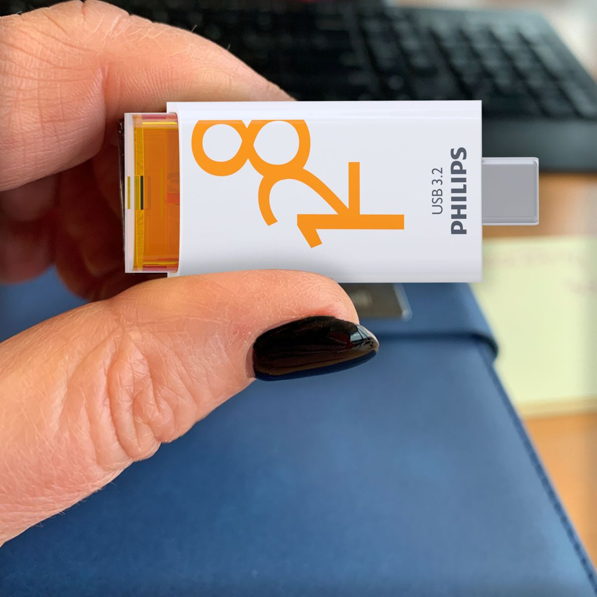 Ręka kobiety trzymająca biało-pomarańczowy pendrive. Na pendrive'ie napis '128' i 'Philips USB 3.2'.