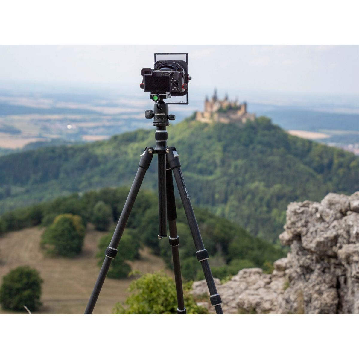Una fotocamera su un treppiede, con un castello sullo sfondo su una collina. Alberi verdi circondano la collina.