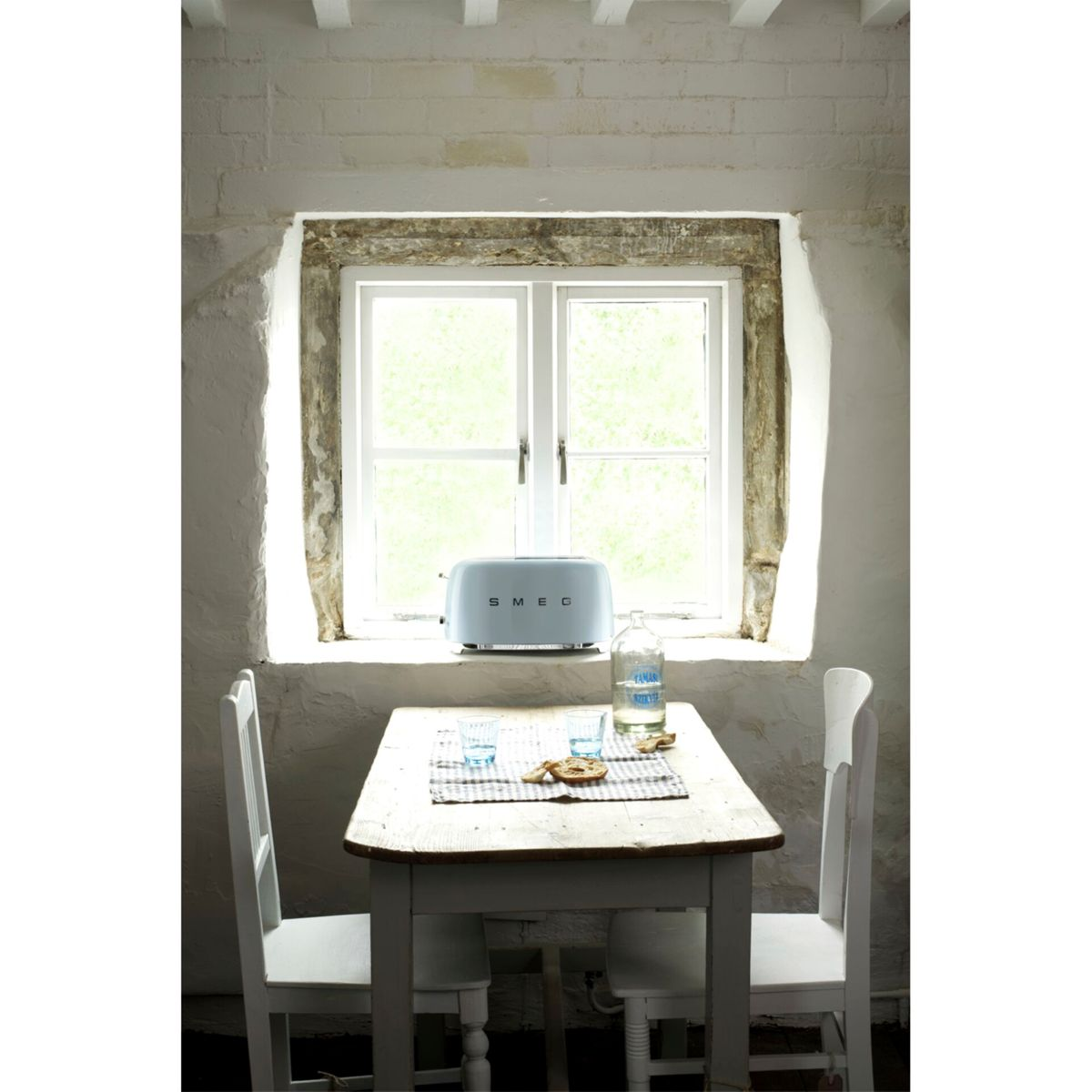 Light blue Smeg toaster sits in a window. A bottle and glasses are on the table with two chairs.