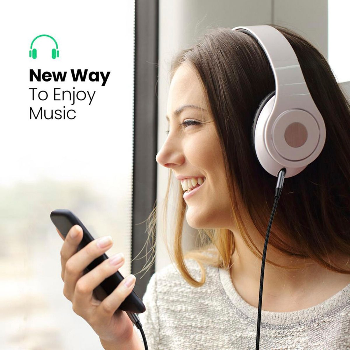 Kobieta uśmiecha się, nosząc słuchawki i trzymając telefon, słuchając muzyki przy oknie.