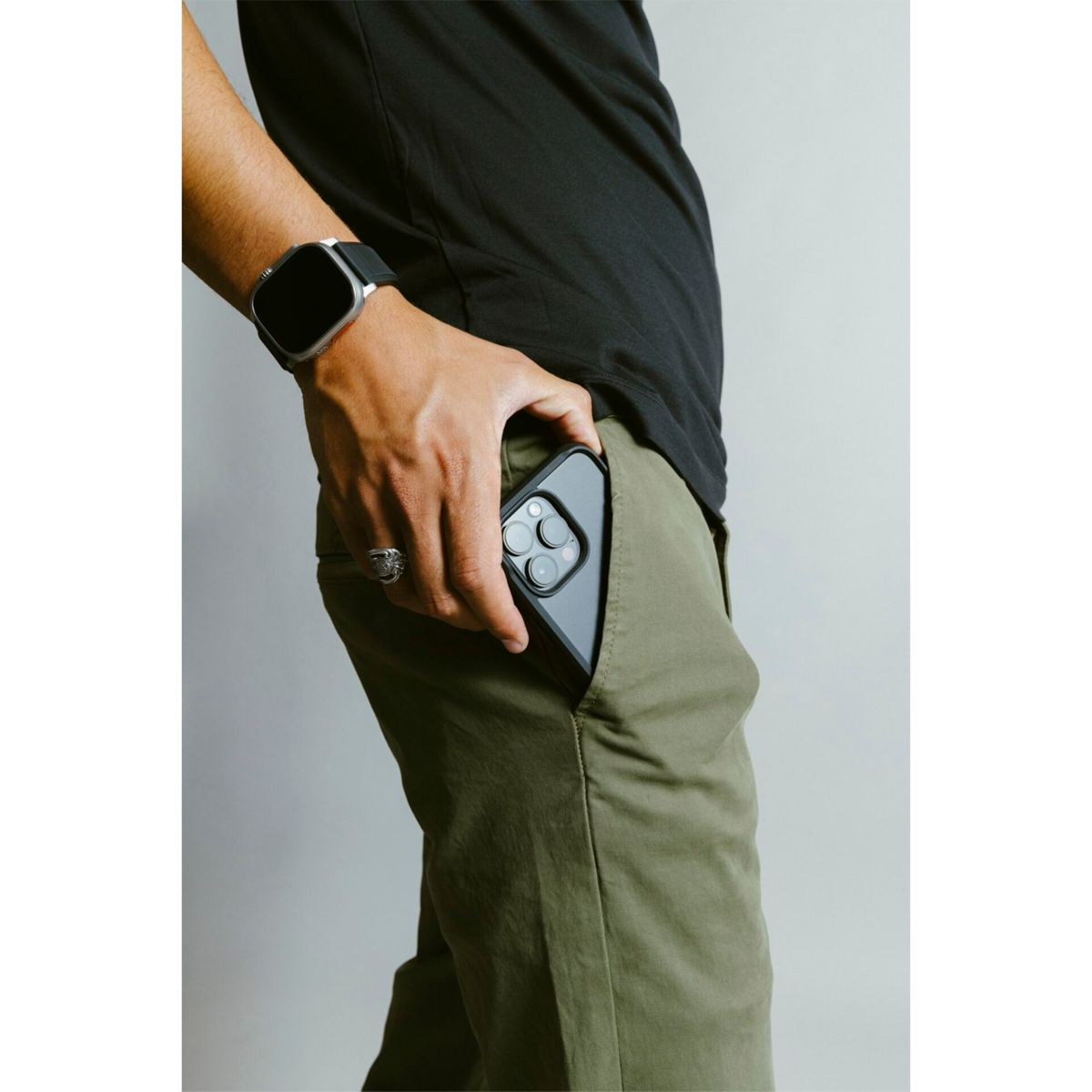 Die Hand einer Person hält ein Telefon in der Tasche. Am Handgelenk befindet sich eine Uhr. Die Hose ist grün.