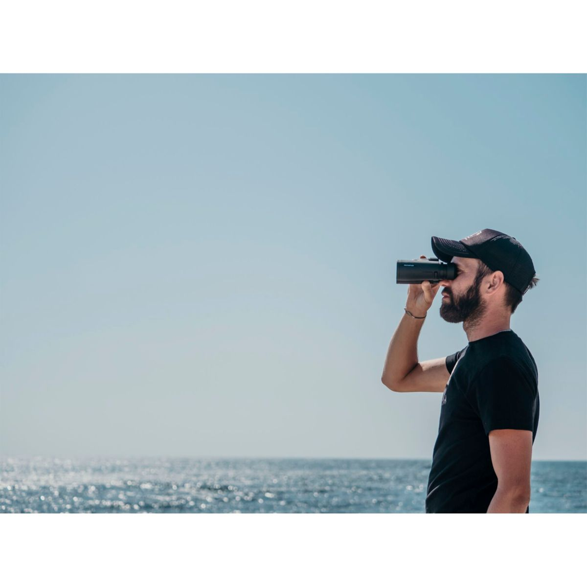 Mężczyzna patrzący przez lornetkę na ocean pod błękitnym niebem.